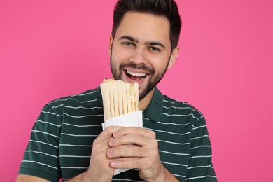 Young man eating tasty shawarma on pink background