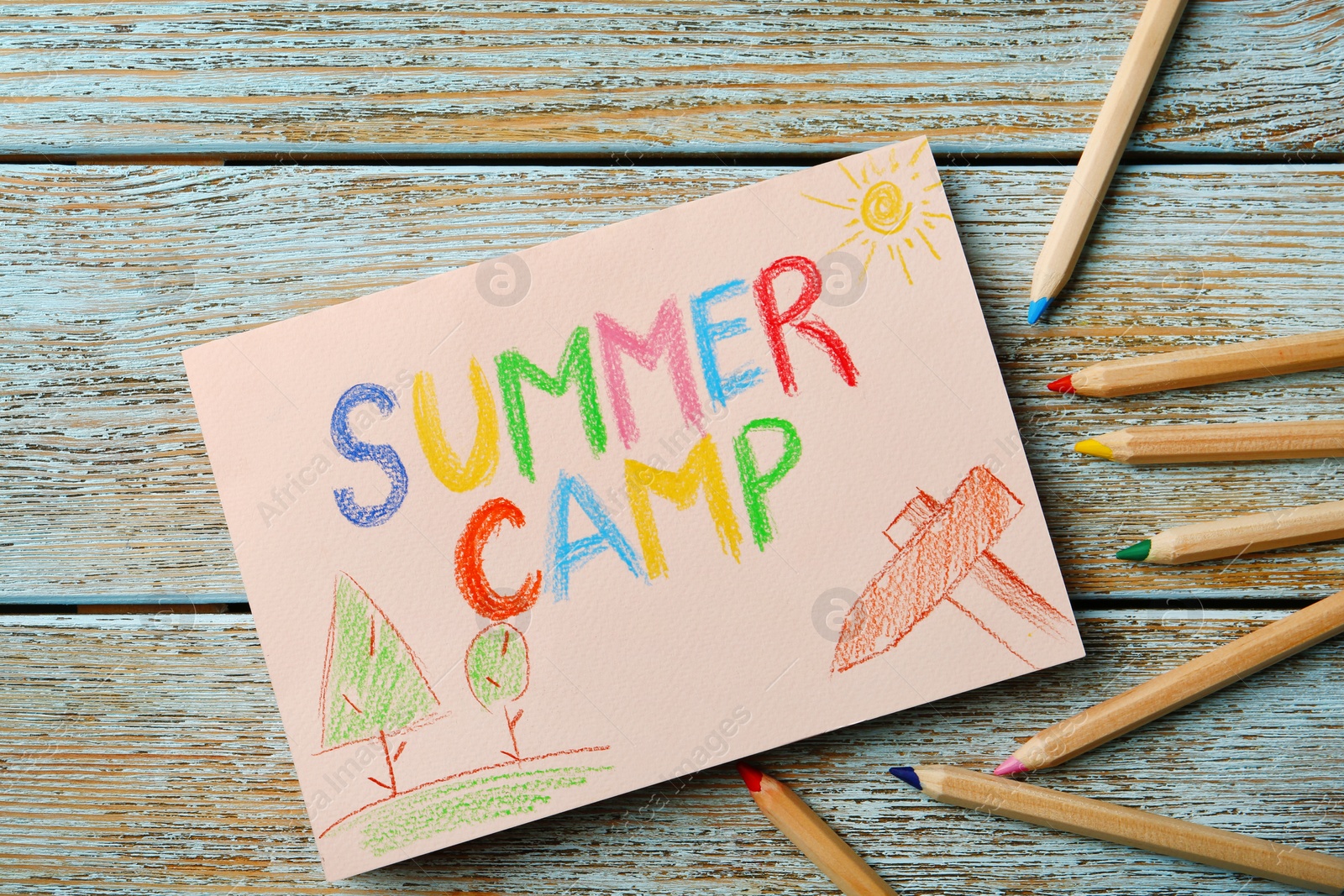 Photo of Paper with written text SUMMER CAMP, drawings and different pencils on wooden background, top view