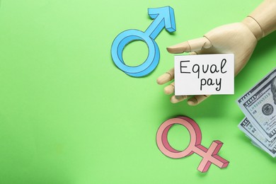 Photo of Equal pay. Wooden mannequin hand, dollar banknotes, paper note, gender symbols on green background, flat lay with space for text