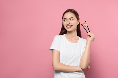 Photo of Beautiful young woman holding sushi roll with chopsticks on pink background. Space for text