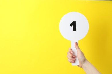Photo of Woman holding auction paddle with number 1 on yellow background, closeup. Space for text