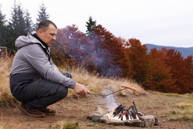 Photo of Man roasting tasty sausage over campfire near forest. Camping season