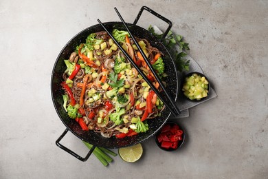 Photo of Stir-fry. Tasty noodles with meat in wok, chopsticks and ingredients on grey textured table, flat lay