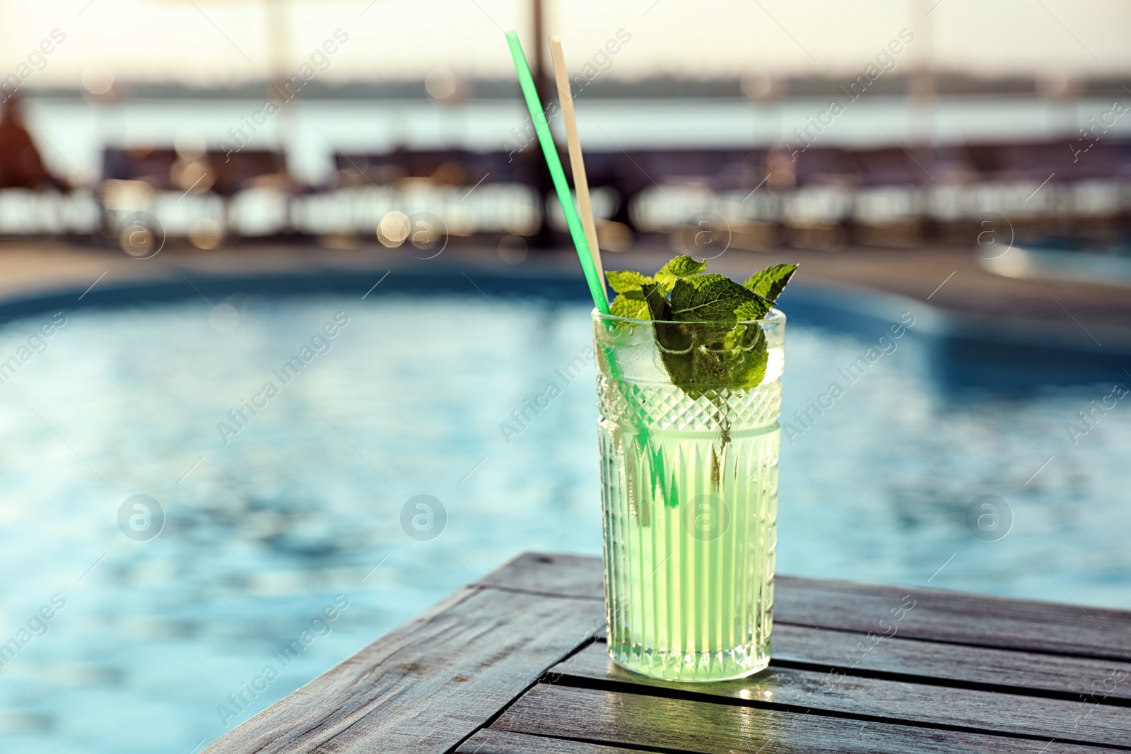 Photo of Glass of fresh summer cocktail on wooden table near swimming pool outdoors. Space for text