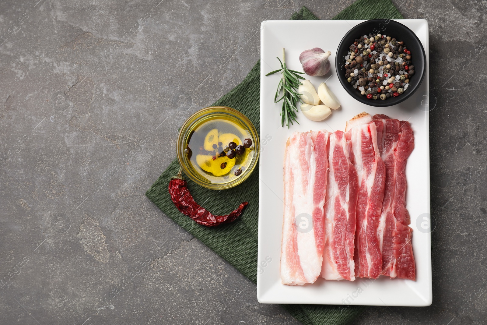 Photo of Pieces of raw pork belly, oil, peppercorns, rosemary and garlic on grey textured table, flat lay. Space for text