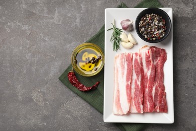 Pieces of raw pork belly, oil, peppercorns, rosemary and garlic on grey textured table, flat lay. Space for text