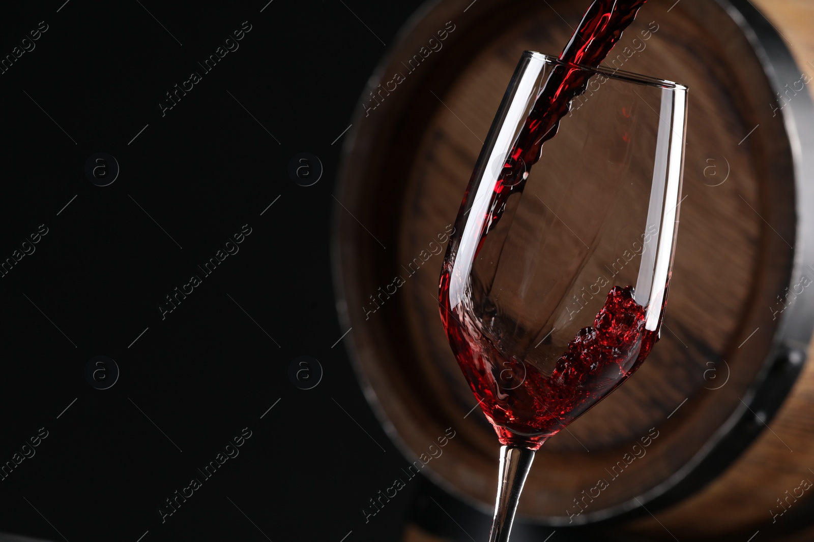 Photo of Pouring red wine into glass near wooden barrel against black background, closeup. Space for text