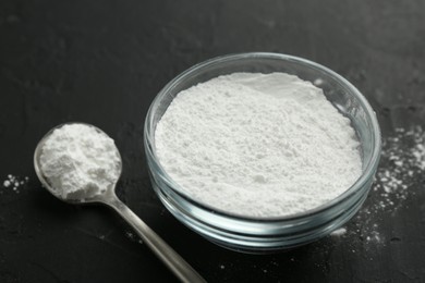 Photo of Baking powder in bowl and spoon on black textured table, closeup