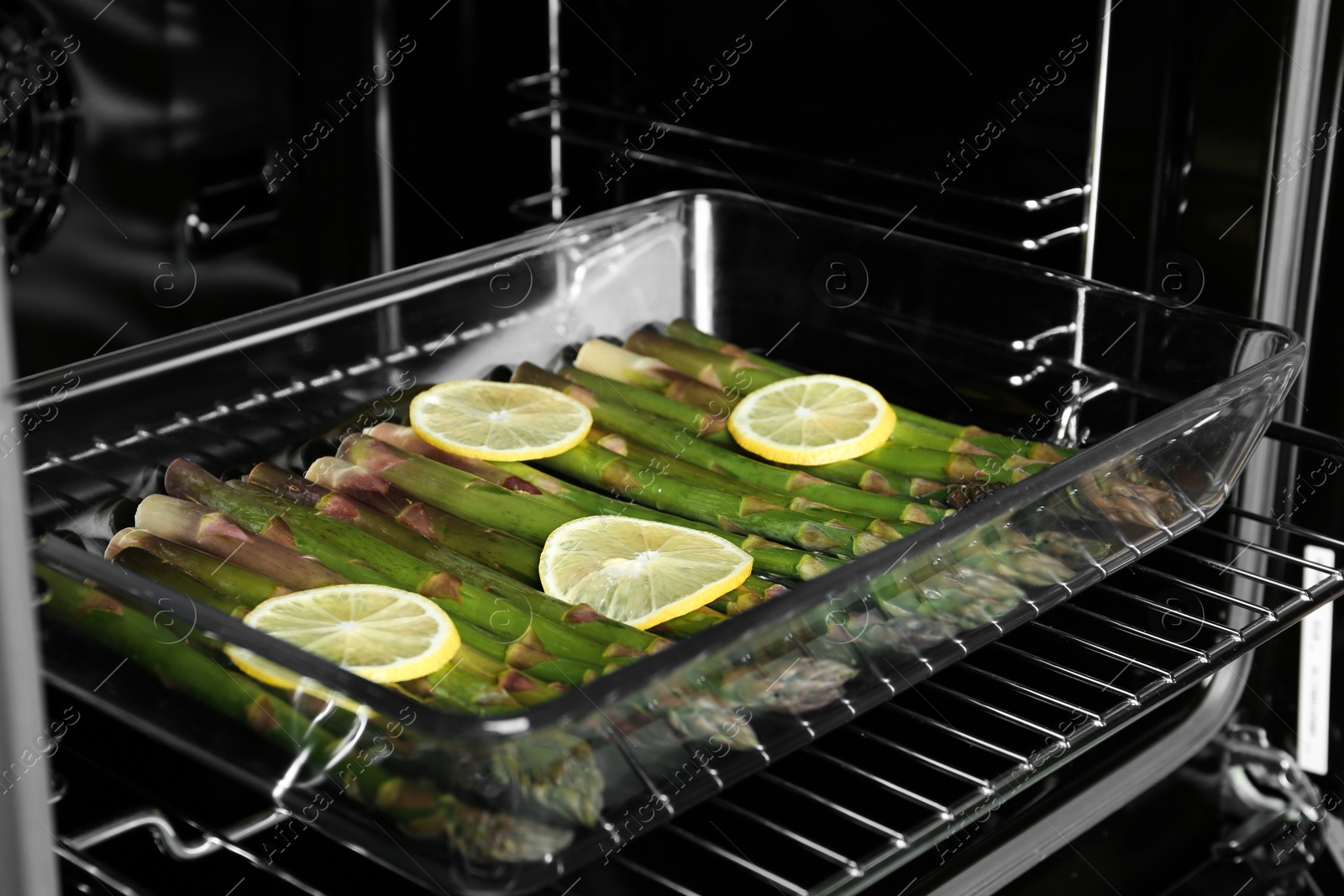 Photo of Raw asparagus with lemon slices in glass baking dish on oven rack, closeup