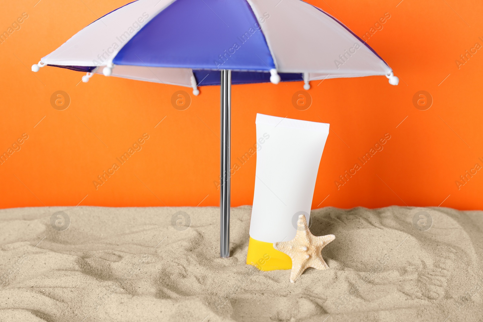 Photo of Suntan product and starfish under beach umbrella on sand against orange background