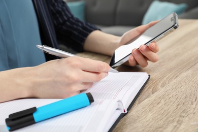 Photo of Woman taking notes while using smartphone at wooden table indoors, closeup