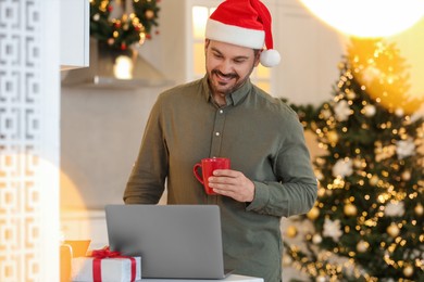Photo of Celebrating Christmas online with exchanged by mail presents. Happy man in Santa hat with cup of drink during video call on laptop at home