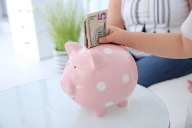 Little girl putting money into piggy bank at table, closeup