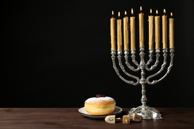 Silver menorah near sufganiyah and dreidels with symbols Nun, He, Pe, Gimel on black background. Space for text