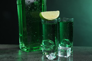 Photo of Absinthe in shot glasses with lime and ice cubes on gray textured table, closeup. Alcoholic drink