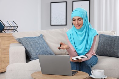 Photo of Muslim woman using video chat on laptop at couch in room. Space for text