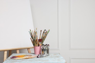 Photo of Easel with blank canvas and different art supplies on wooden table near white wall. Space for text