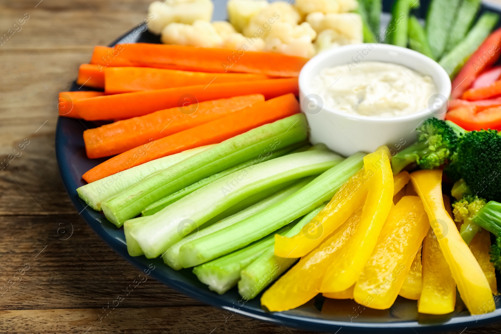 Photo of Plate with celery sticks, other vegetables and dip sauce on wooden table, closeup