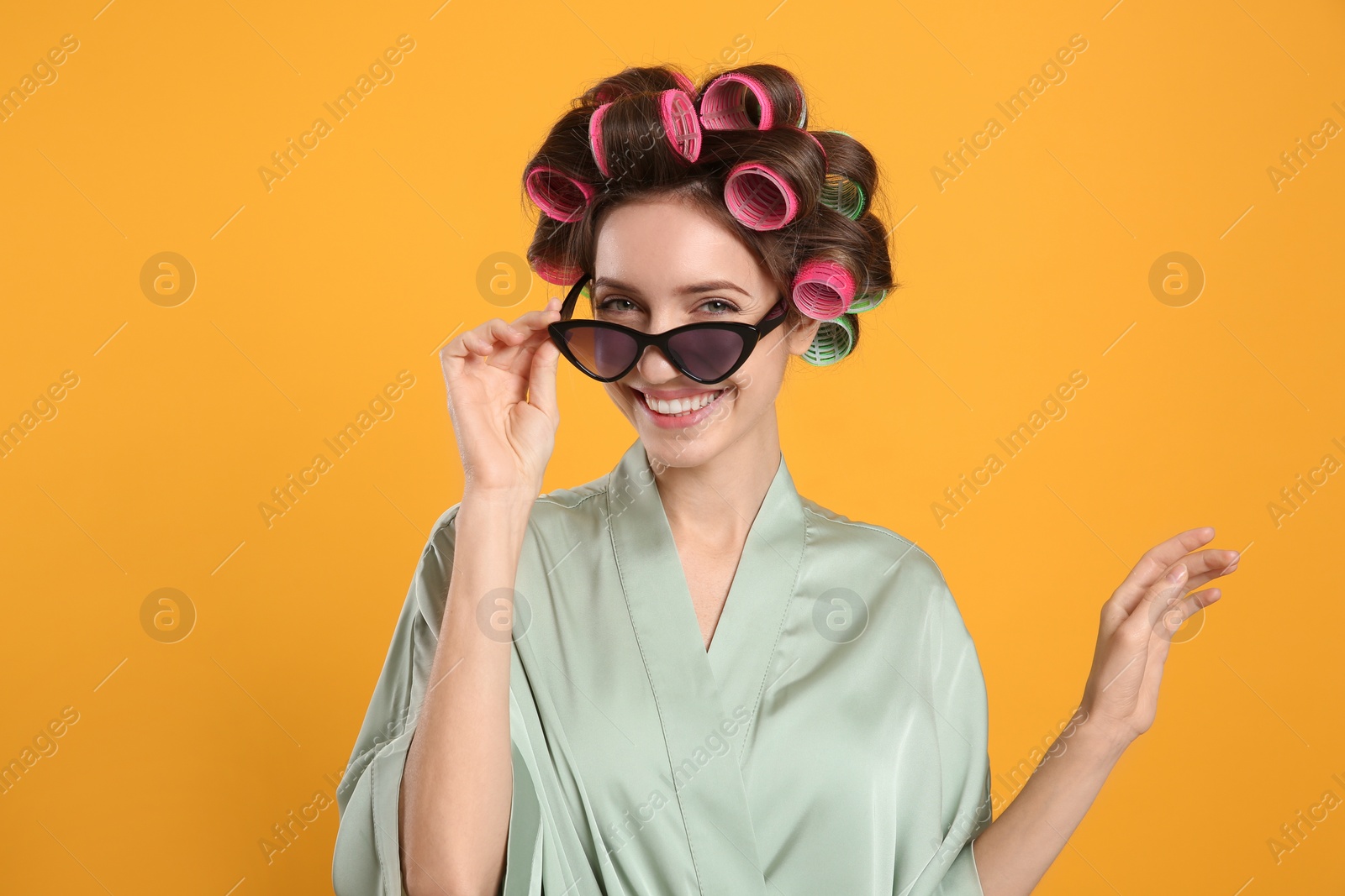 Photo of Beautiful young woman in silk bathrobe with hair curlers and sunglasses on yellow background