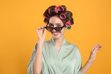 Beautiful young woman in silk bathrobe with hair curlers and sunglasses on yellow background