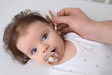 Father cleaning ear of his baby with cotton bud on bed, closeup