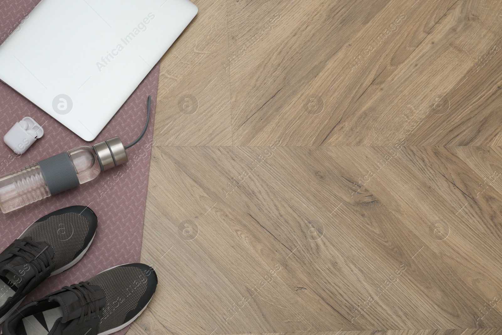 Photo of Exercise mat, laptop, bottle of water, wireless earphones and shoes on wooden floor, flat lay. Space for text