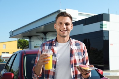 Young man with doughnut and juice near car at gas station