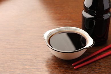 Photo of Bottle, bowl with soy sauce and chopsticks on wooden table, closeup. Space for text