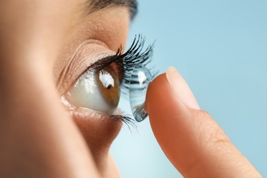 Photo of Young woman putting contact lens in her eye on light blue background, closeup