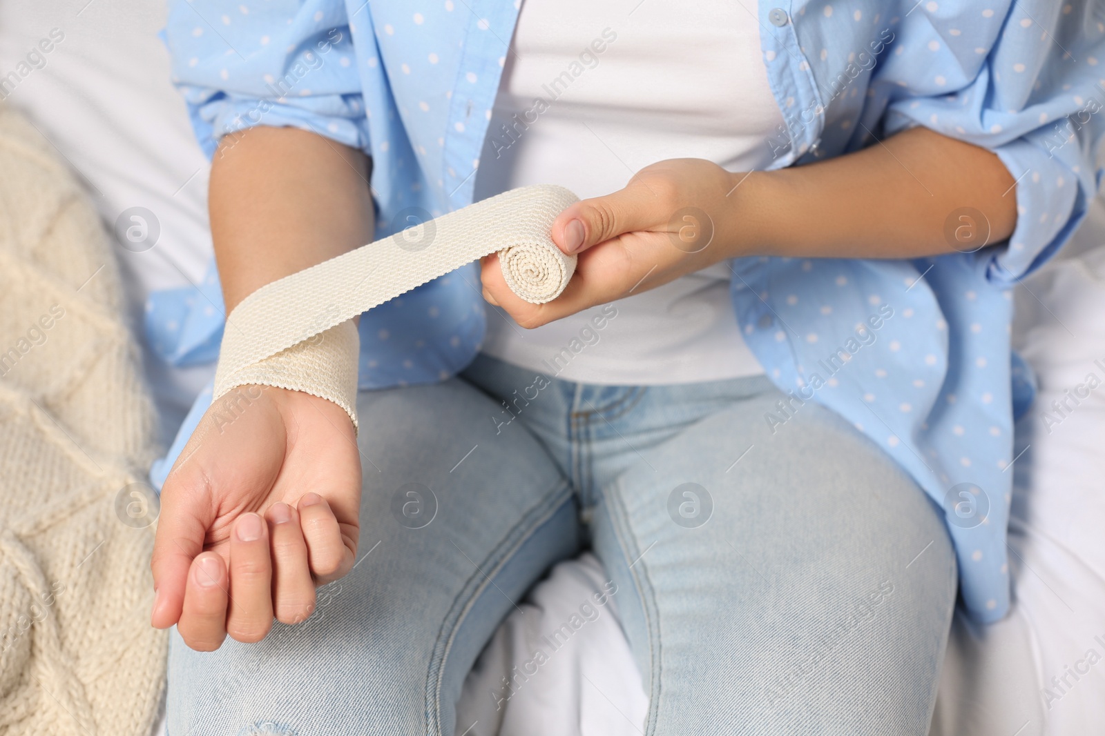 Photo of Young woman applying medical bandage onto wrist on bed, closeup