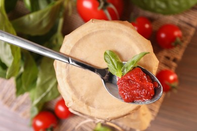 Jar of tasty tomato paste with spoon and basil on table, flat lay