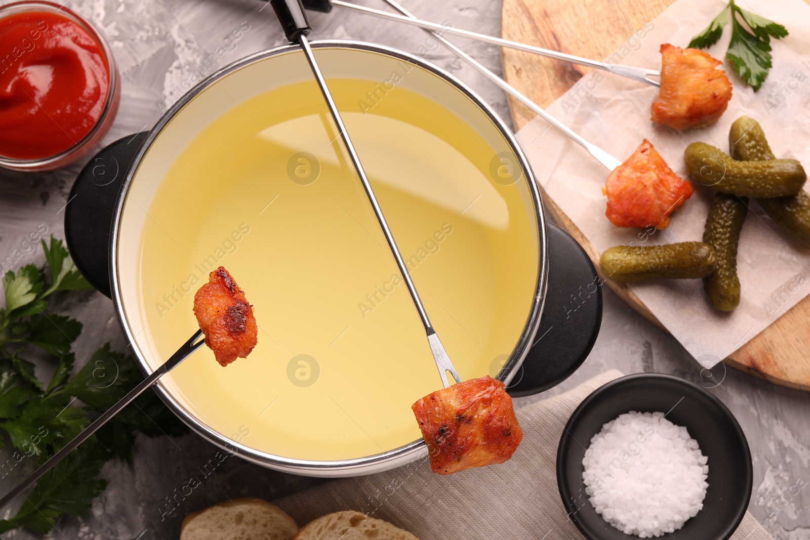 Photo of Fondue pot, forks with fried meat pieces, salt and other products on grey textured table, flat lay