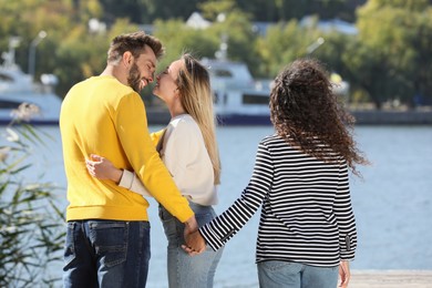 Photo of Man holding hands with another woman behind his girlfriend's back near river on sunny day. Love triangle