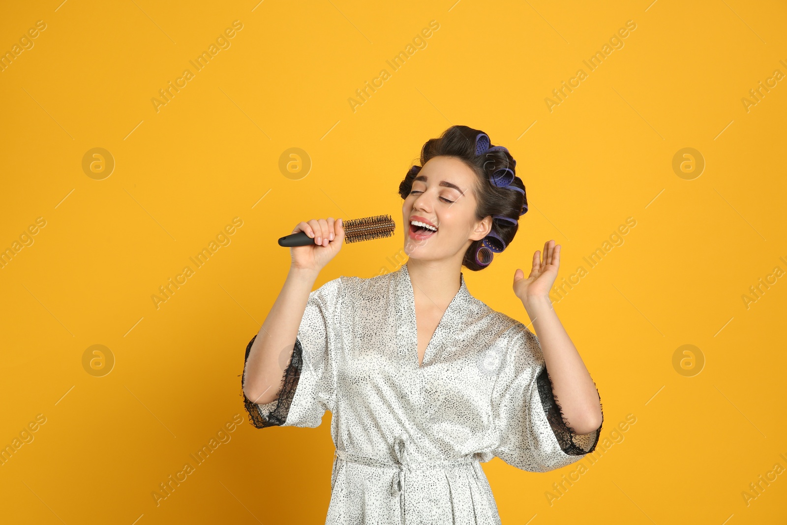 Photo of Happy young woman in silk bathrobe with hair curlers singing into hairbrush on orange background