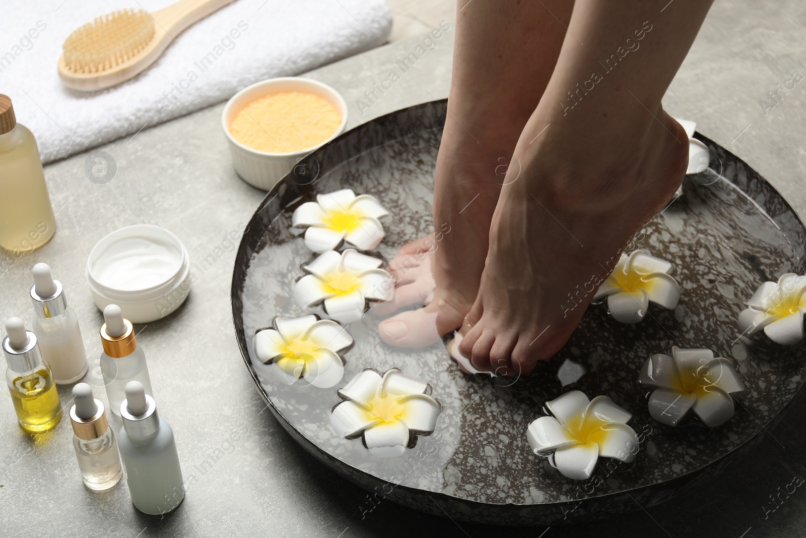 Photo of Woman soaking her feet in bowl with water and flowers on light grey floor, closeup. Spa treatment