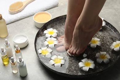 Woman soaking her feet in bowl with water and flowers on light grey floor, closeup. Spa treatment