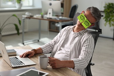 Man with fake eyes painted on sticky notes snoozing at workplace in office