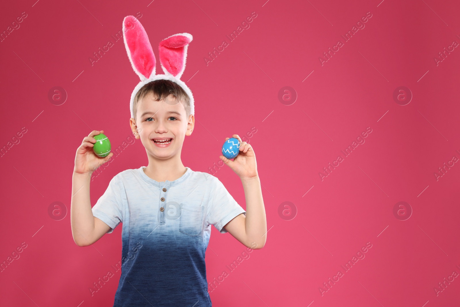 Photo of Little boy in bunny ears headband holding Easter eggs on color background, space for text