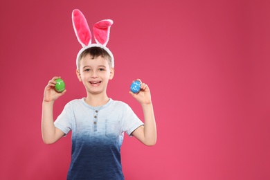 Little boy in bunny ears headband holding Easter eggs on color background, space for text