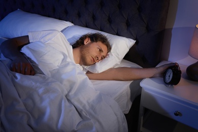 Photo of Sleepy young man turning off alarm clock on nightstand. Bedtime