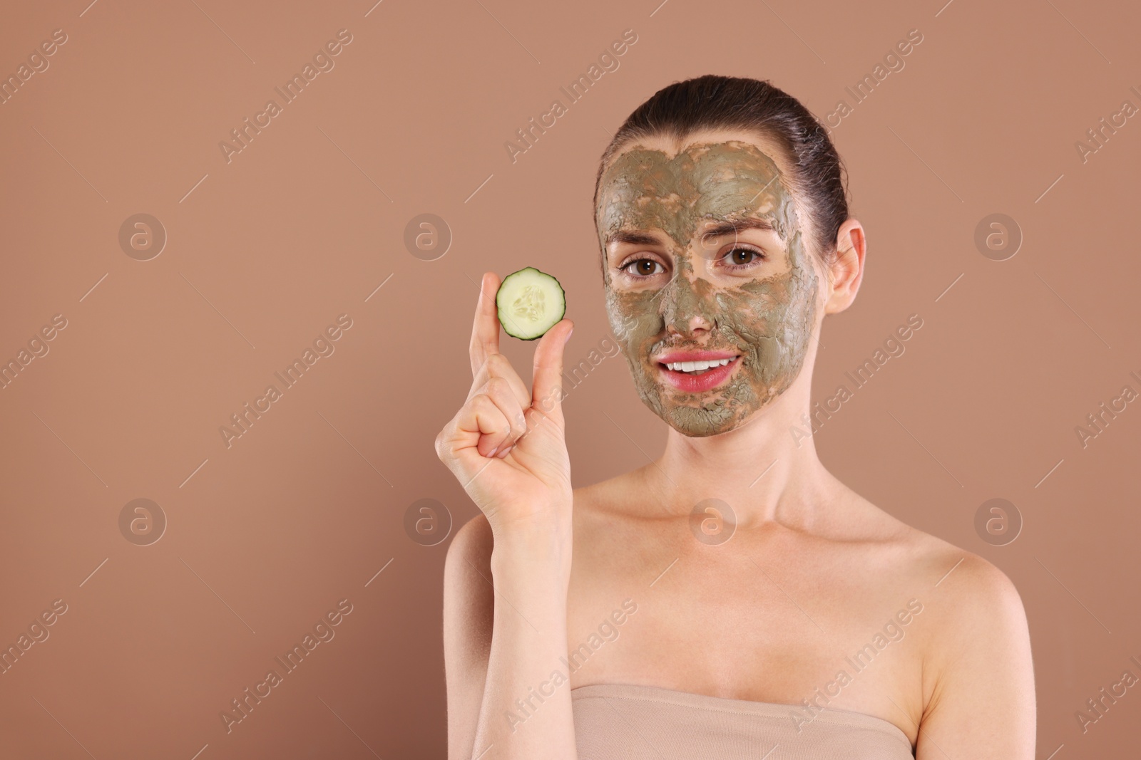 Photo of Beautiful woman with clay mask holding piece of cucumber on light brown background, space for text