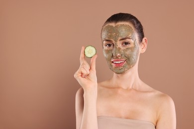 Beautiful woman with clay mask holding piece of cucumber on light brown background, space for text