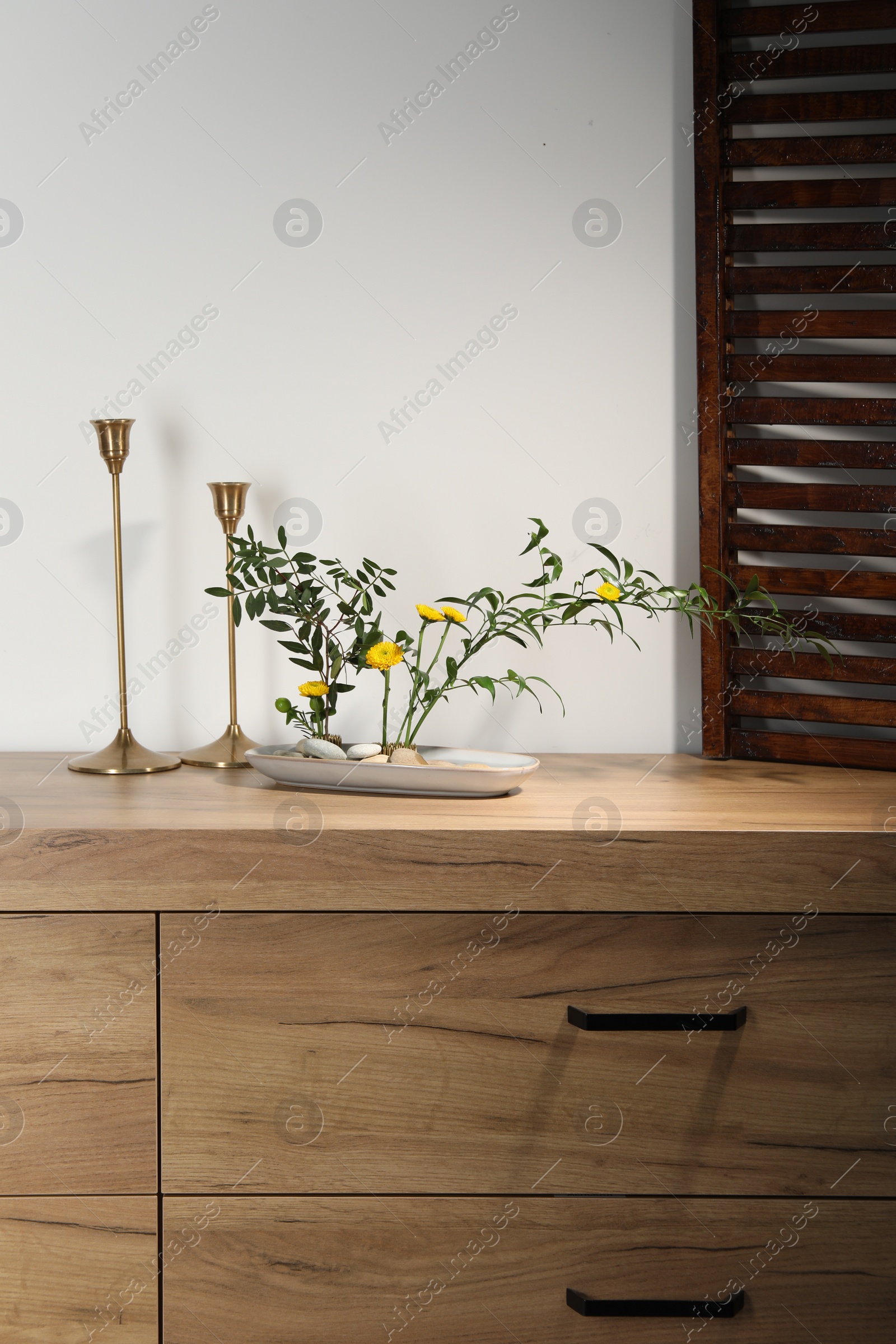 Photo of Stylish ikebana with beautiful yellow flowers, green branches and candlesticks carrying cozy atmosphere at home