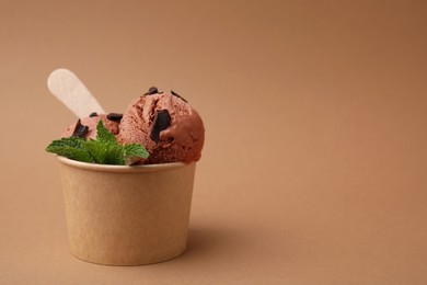 Photo of Paper cup with tasty chocolate ice cream, sticks and mint leaves on light brown background. Space for text