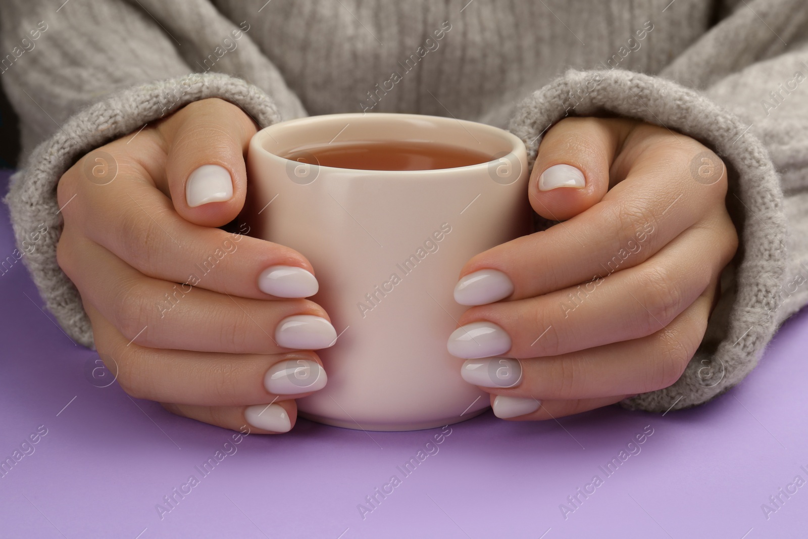 Photo of Woman with white polish on nails holding cup of hot drink on violet background, closeup