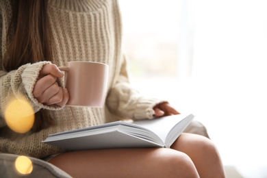 Photo of Young woman with cup of coffee reading book near window at home, closeup