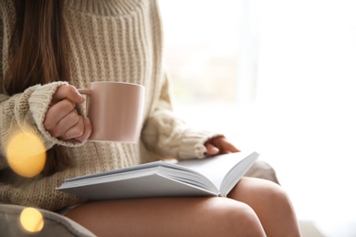 Young woman with cup of coffee reading book near window at home, closeup