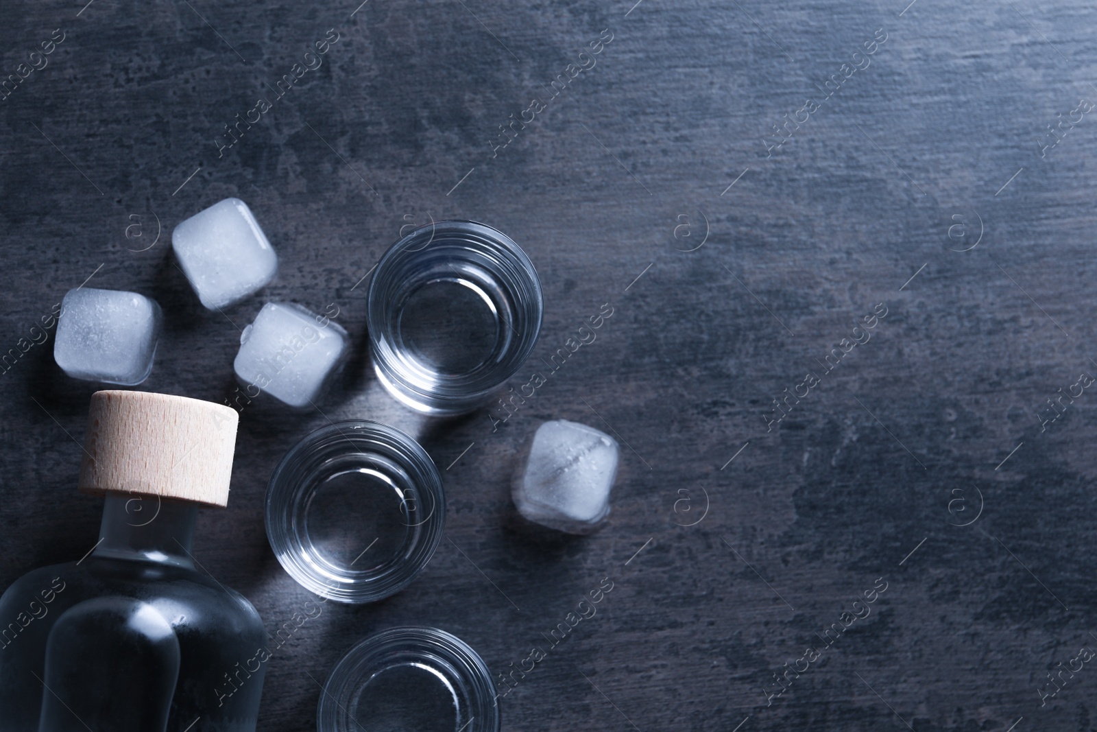 Photo of Bottle of vodka and shot glasses with ice on dark table, flat lay. Space for text