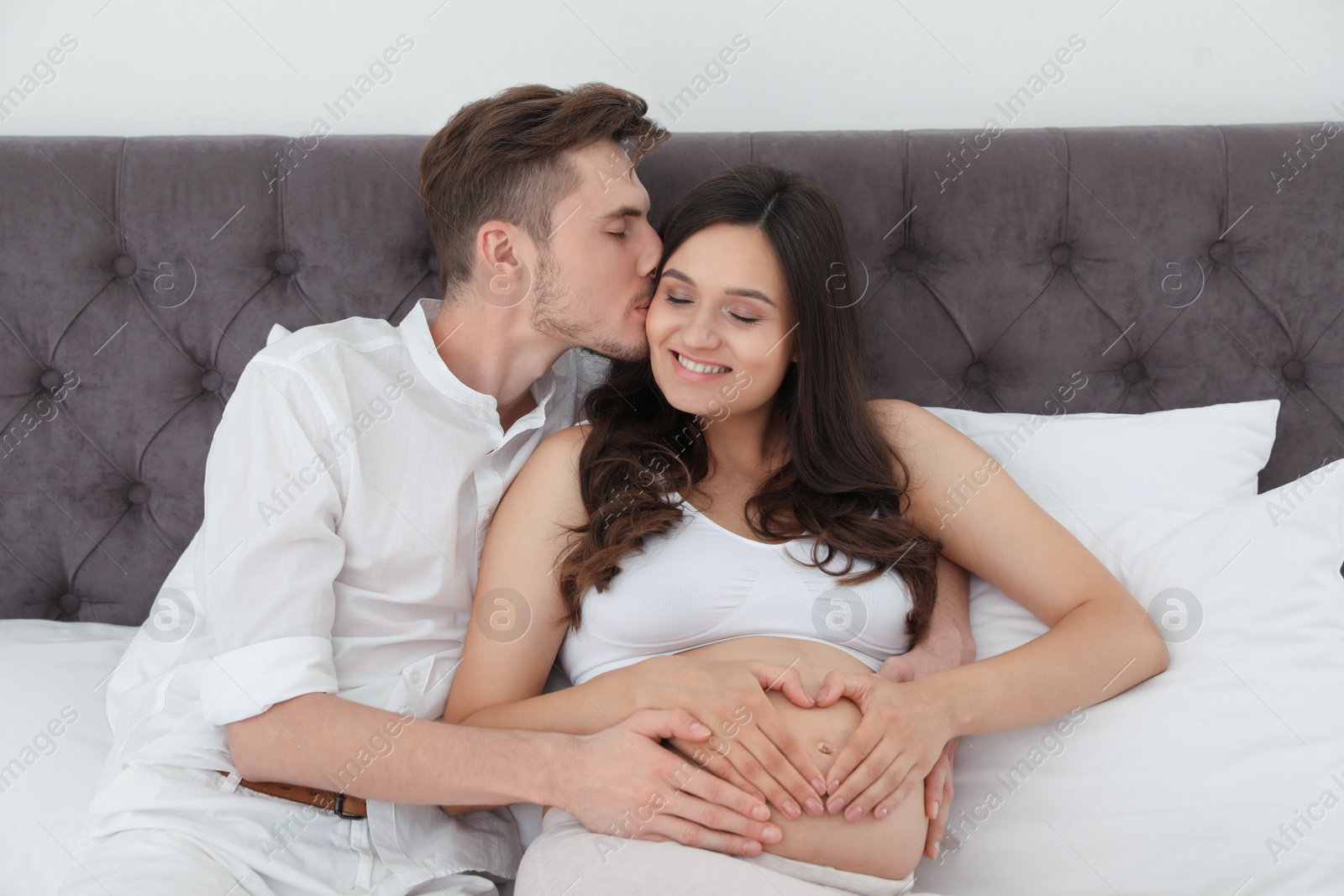 Photo of Young husband and his pregnant wife showing heart with hands in bed