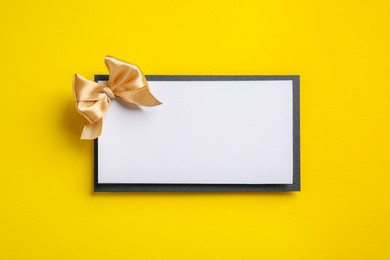 Photo of Blank gift card with golden bow on yellow background, top view